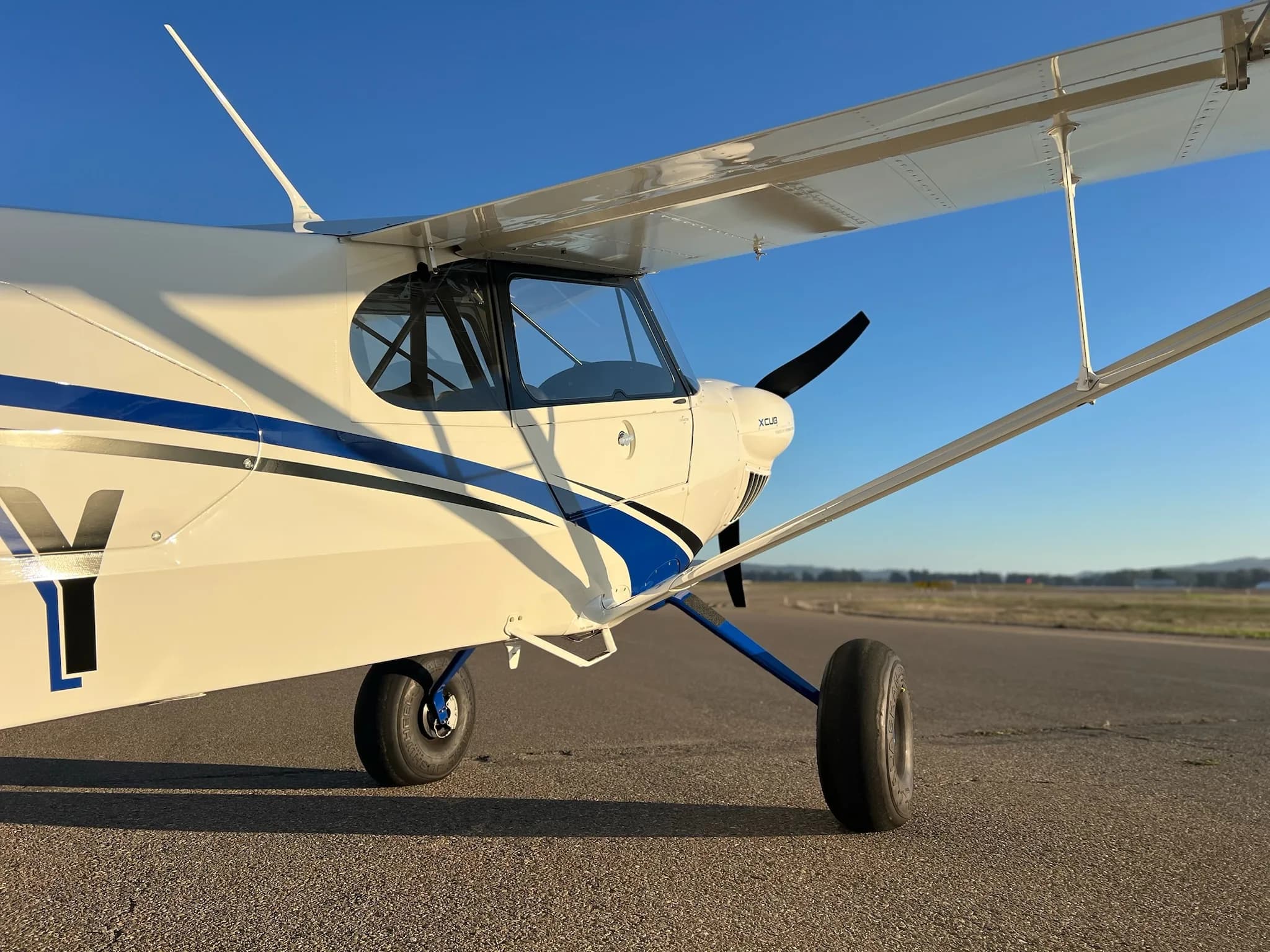 A close-up view of the front-right side of a white X-Cub aircraft with blue accents, showing the propeller and landing gear.