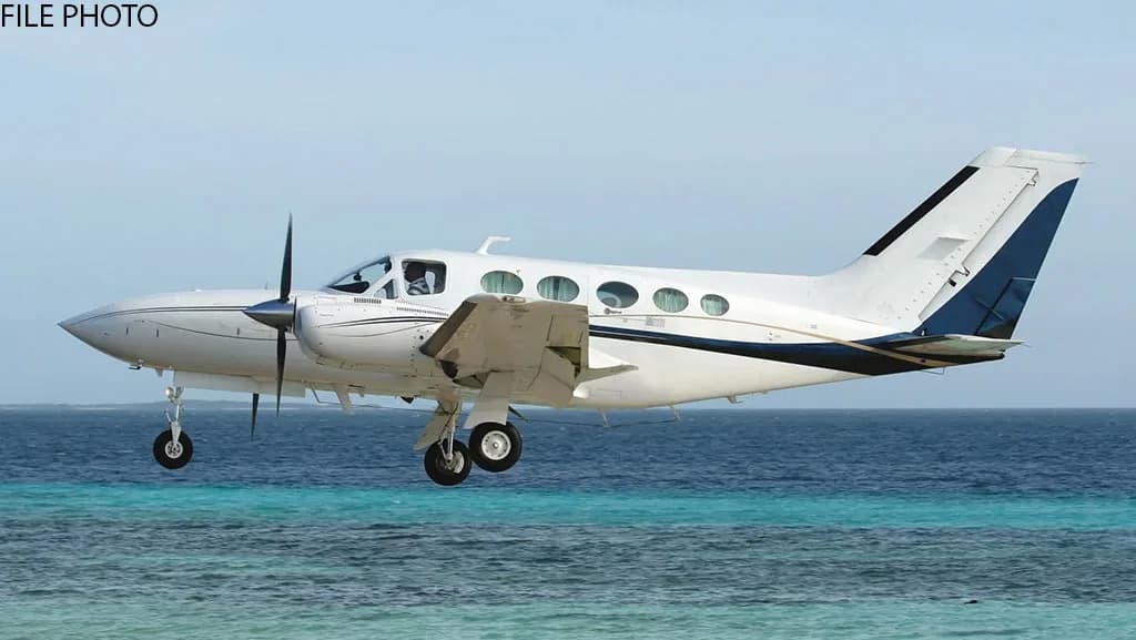 A white twin-engine propeller aircraft with blue and black accents flies low over clear blue tropical water, with its landing gear extended.