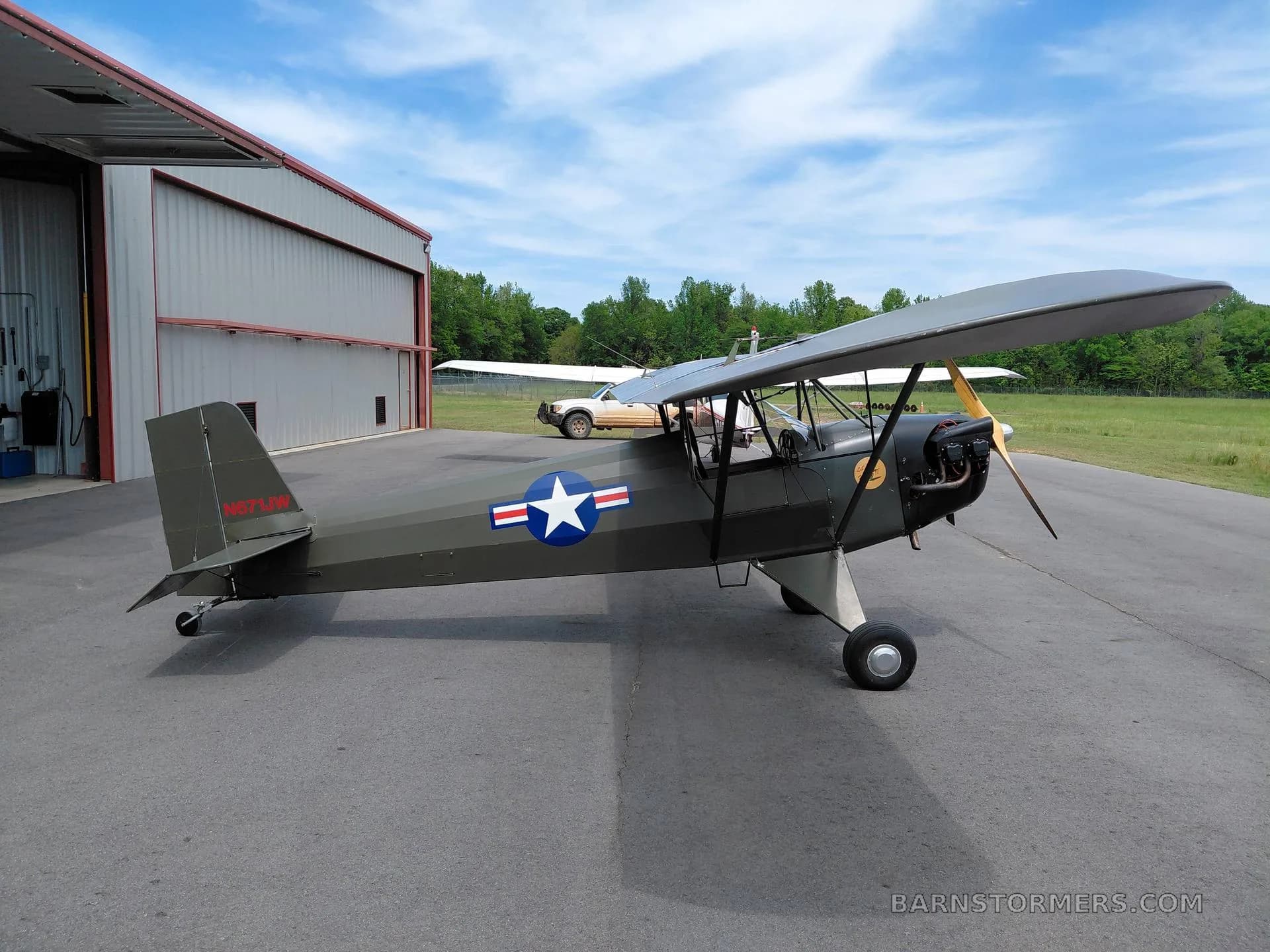 A dark green single-engine, high-wing aircraft with a US Army star insignia, parked on an asphalt apron with hangars and trees in the background.