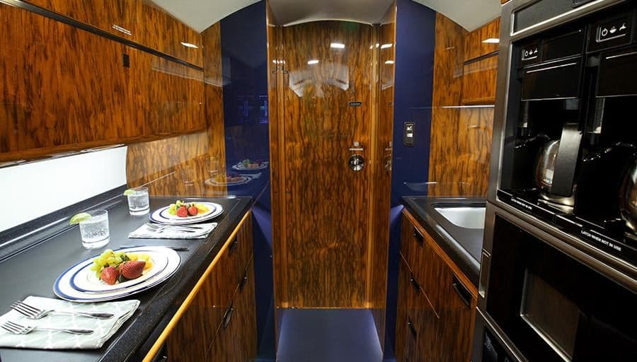 The galley area of the private jet, featuring a countertop with plates, a sink, and built-in appliances with dark wood cabinetry.