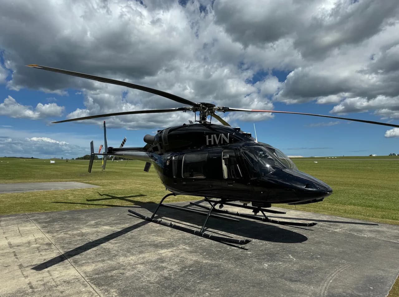 A black helicopter with 'HVN' registration is parked on a concrete pad at an airfield under a partly cloudy sky.