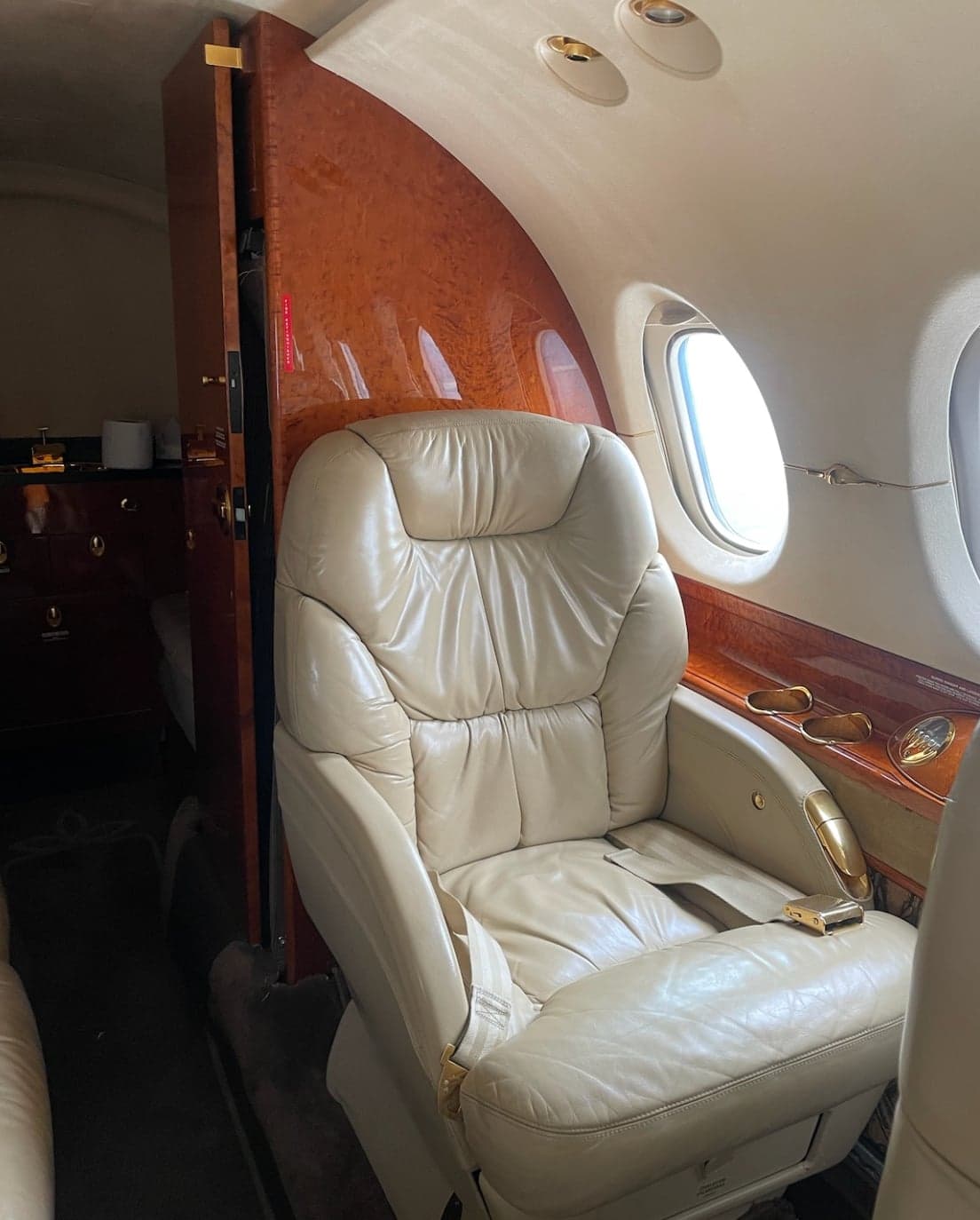 A single beige leather seat in the luxurious interior of a business jet, with wood paneling and a window.