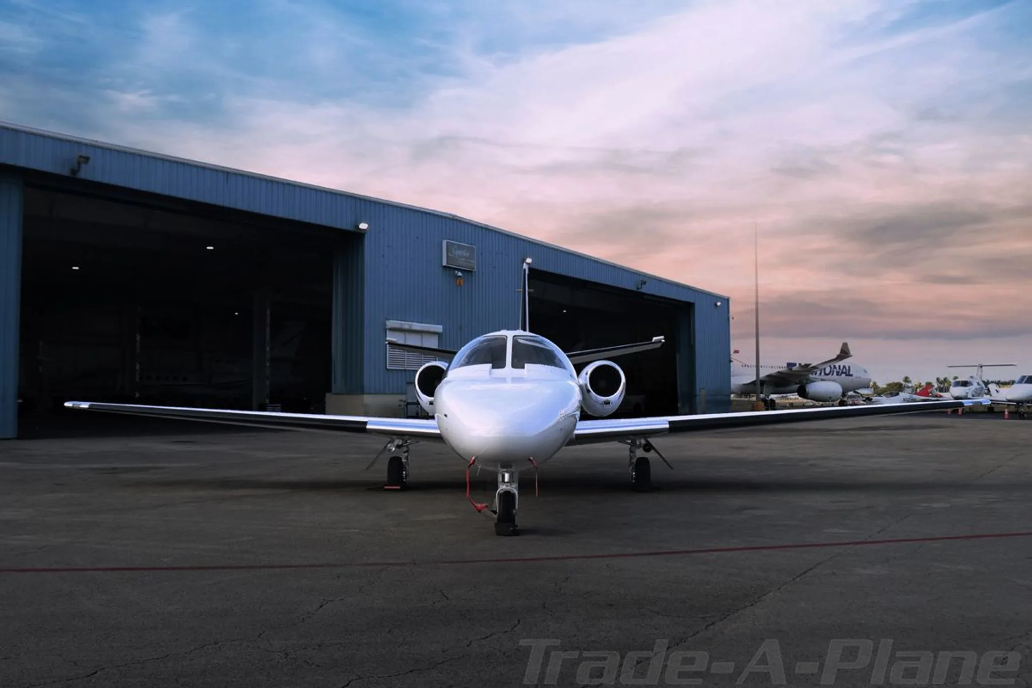 A front view of a white Cessna CitationJet N1EJ parked on an tarmac in front of a blue hangar at sunset.