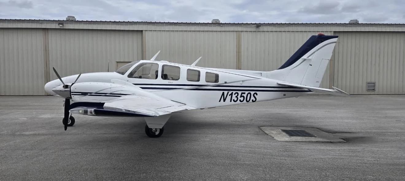 A white and blue Beechcraft Baron N1350S parked on an asphalt apron in front of a hangar.