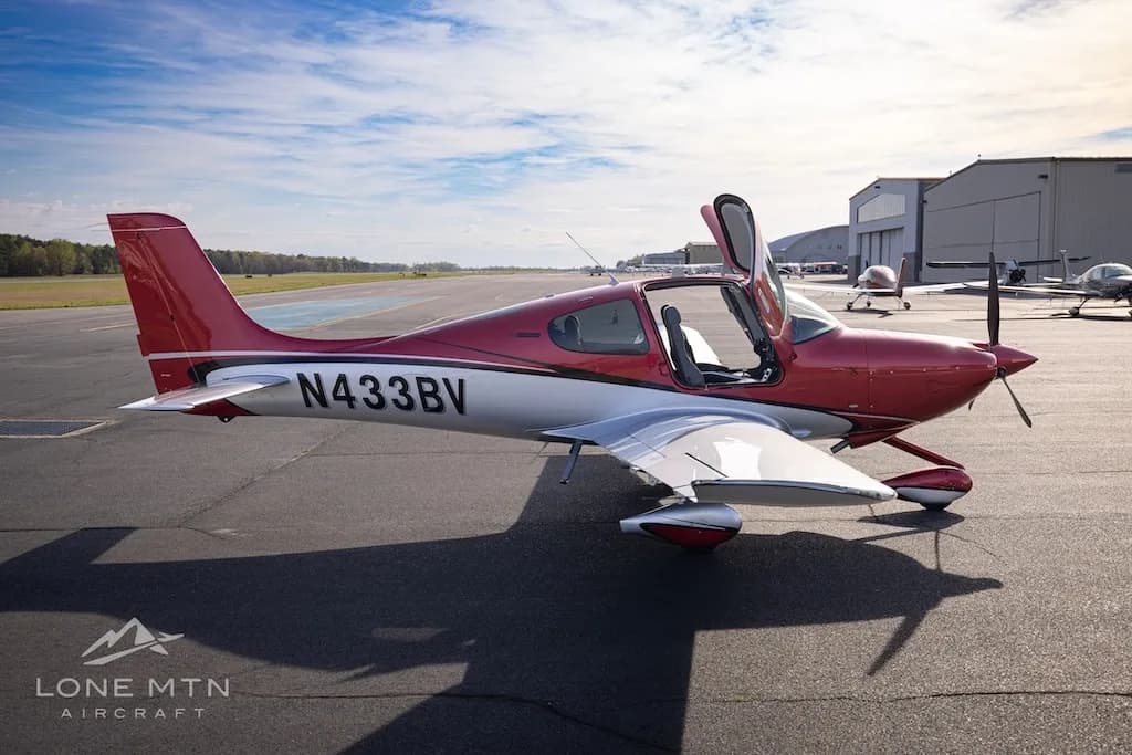 A side view of the red and silver Cirrus SR22 aircraft N433BV with its doors open, parked on an airfield with other aircraft in the distance.