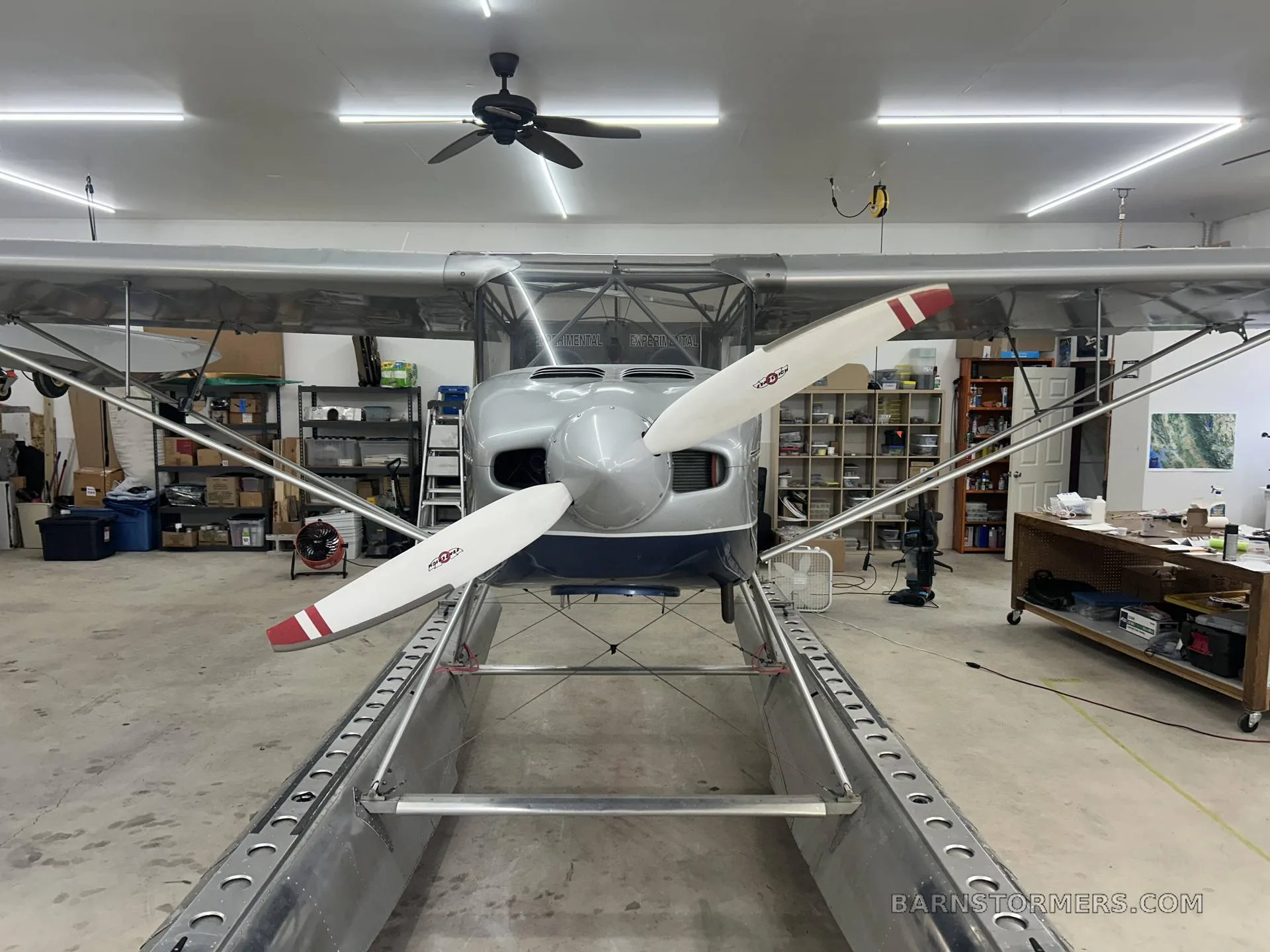 A silver and blue floatplane with a two-bladed propeller is viewed head-on inside a hangar.