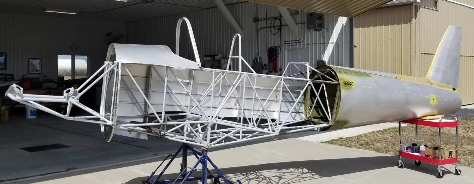 The skeletal frame of an aircraft fuselage, with the tail section attached, on stands outside a hangar.