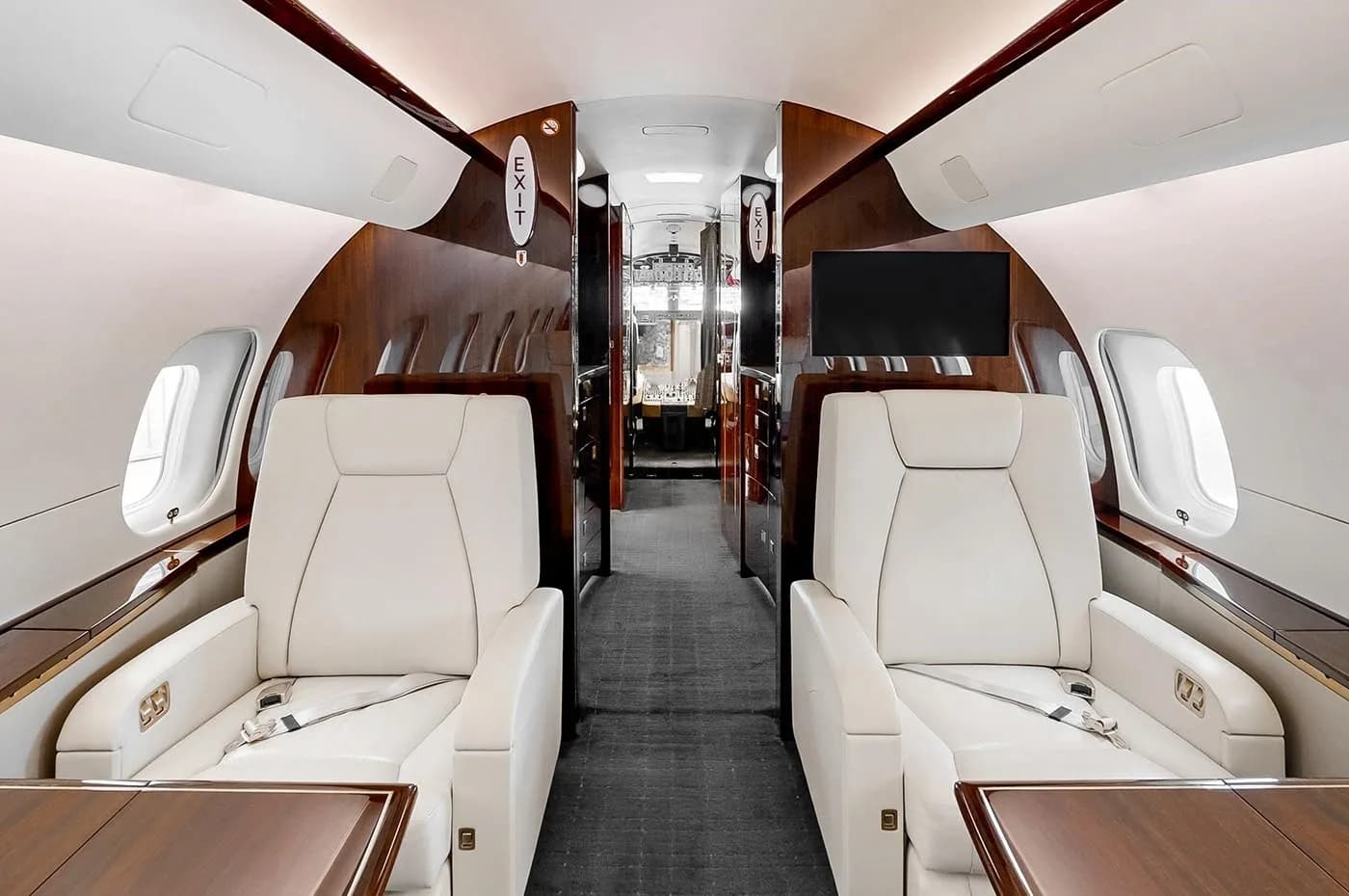 A view down the aisle of a business jet's cabin, showing two cream leather seats with tables and rich dark wood paneling.