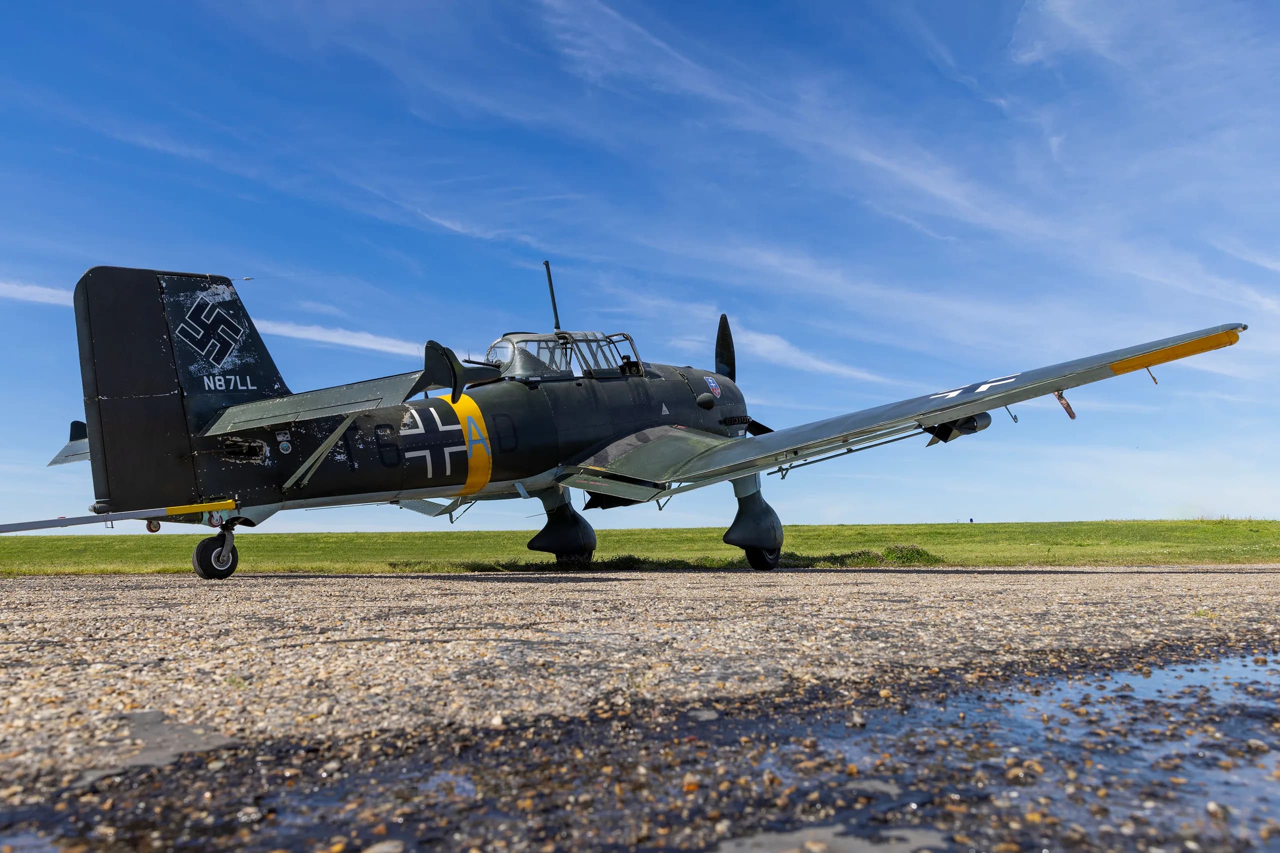 A rear-side view of a dark green Junkers Ju 87 Stuka with tail number N87LL, parked on an airfield.