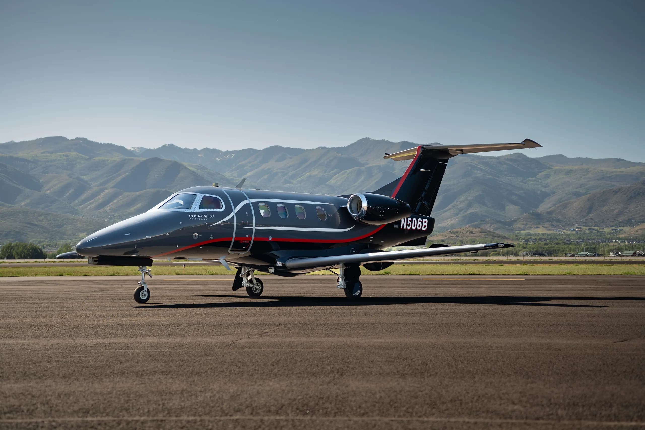 A dark gray and black Embraer Phenom 100 with red and white stripes, registration N506B, parked on a runway during the day with mountains in the background.