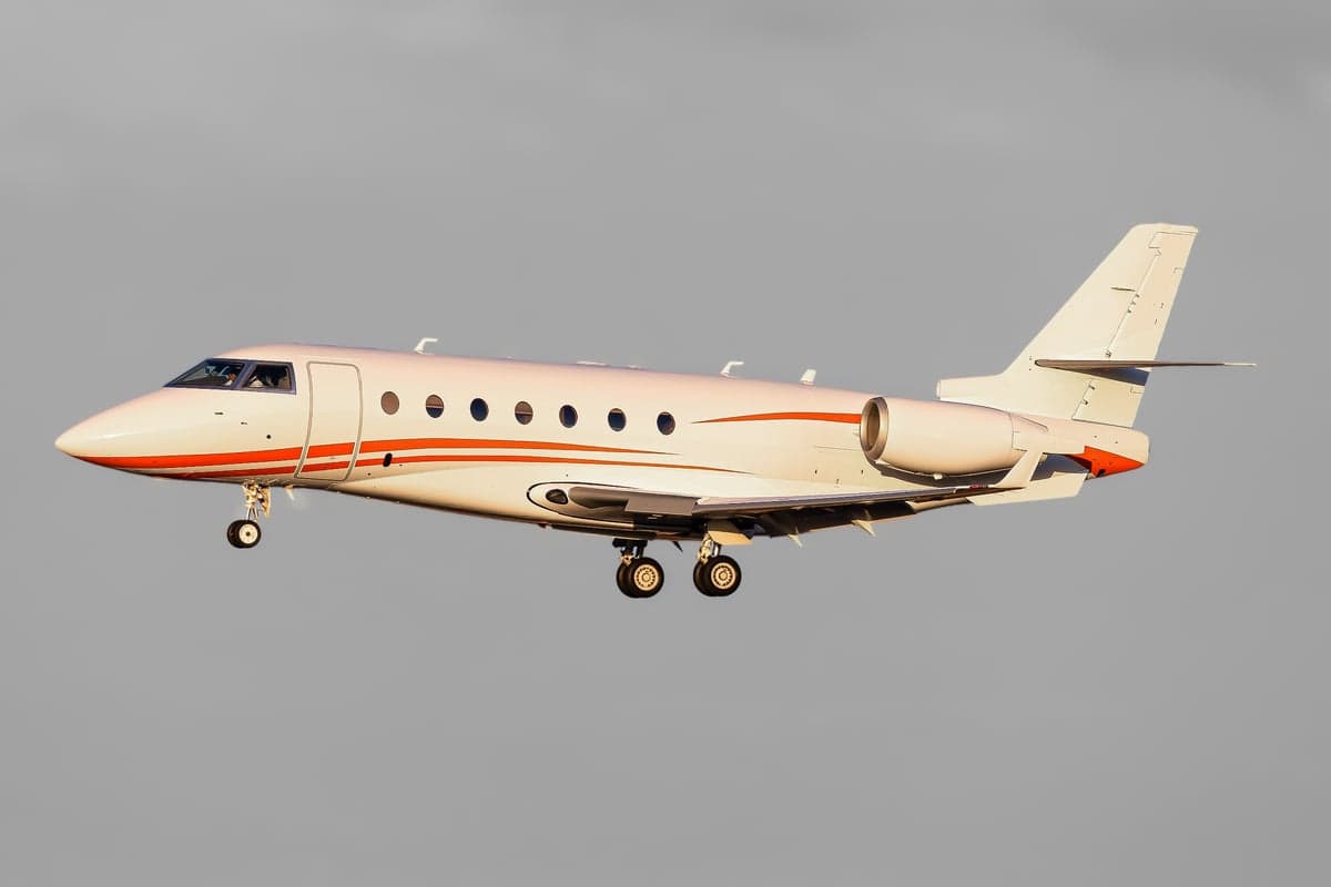 A white business jet with orange stripes on its fuselage and tail, flying against a grey sky with landing gear extended.