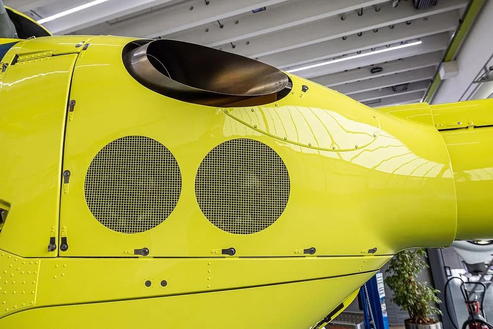 A close-up of the bright yellow rear fuselage of an Airbus H135 helicopter, showing an exhaust port and two circular vents.