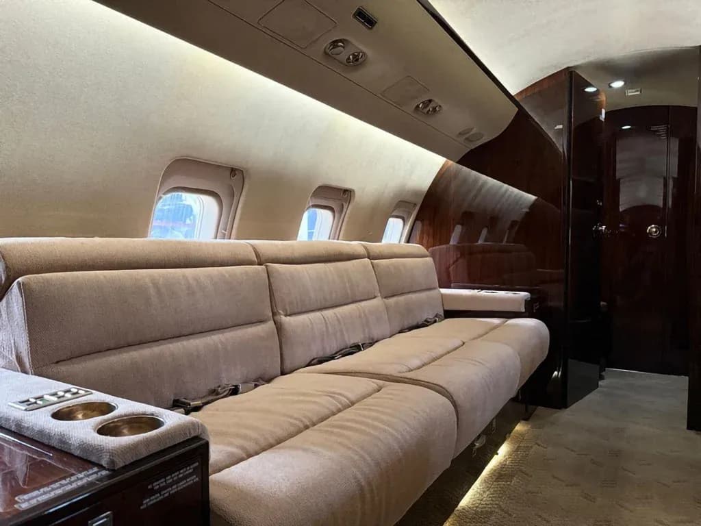 A long beige sofa with cup holders is visible in the private jet cabin, with dark wood accents and windows.