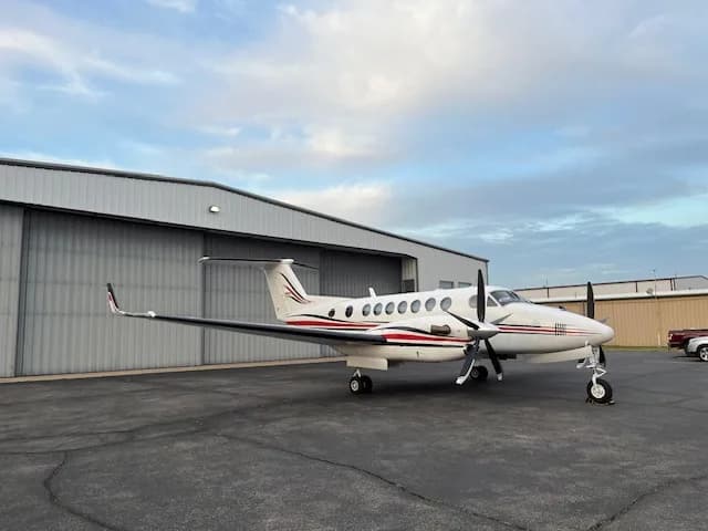 A white Beechcraft King Air with red and black stripes parked on an tarmac in front of a hangar, viewed from the right side.