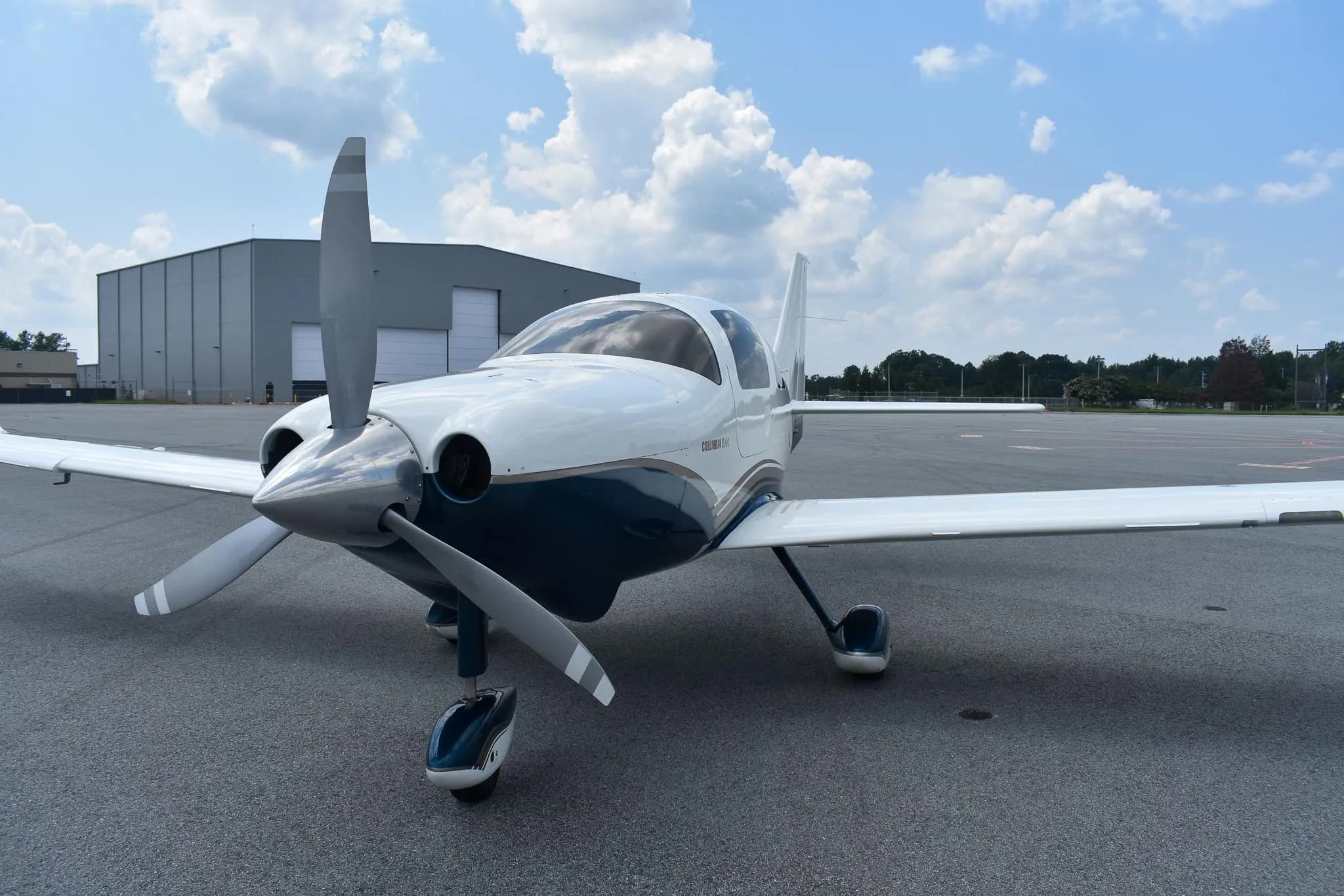 A front view of the white and blue Columbia 400 aircraft, registration N371BG, parked on an airfield with a hangar in the background.