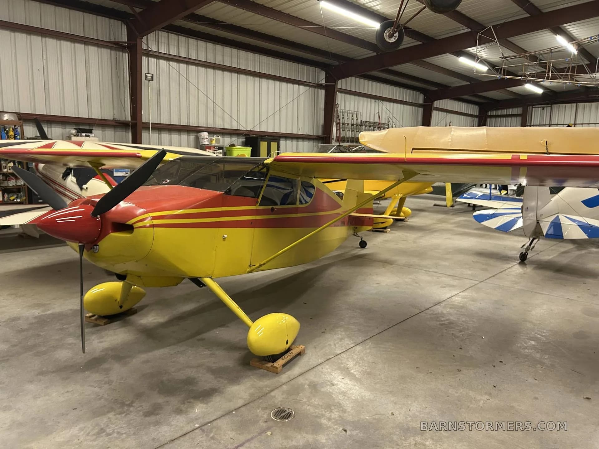 A yellow and red single-engine aircraft is parked in a hangar, viewed from the front-left.