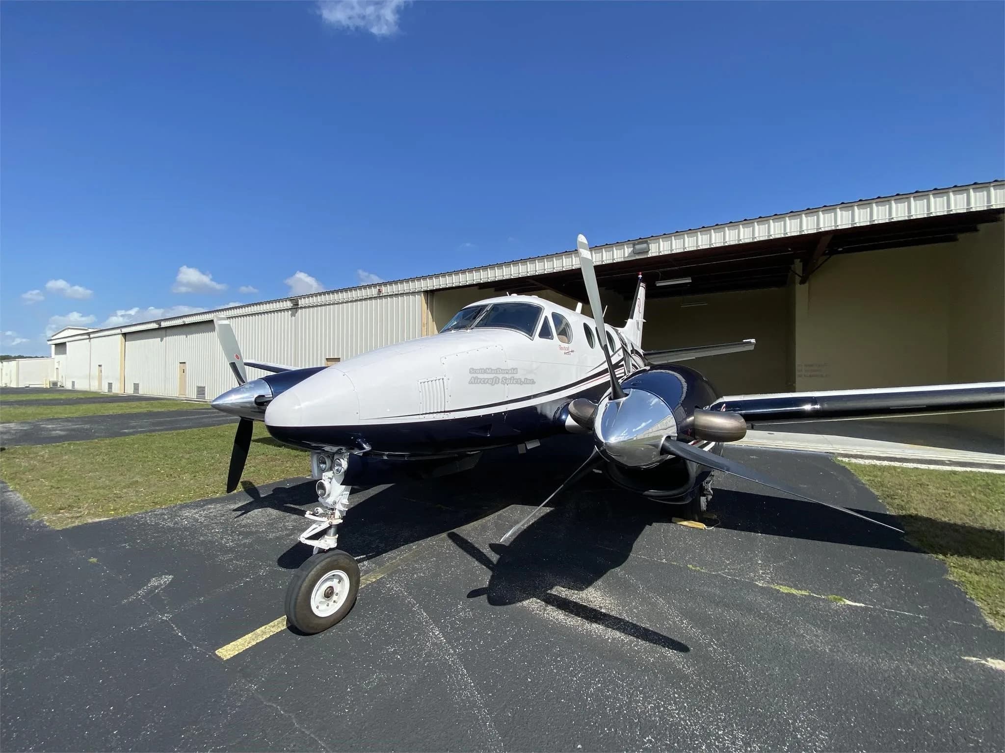 A head-on view of a white and dark blue Beechcraft King Air with its propellers, parked on an asphalt apron.