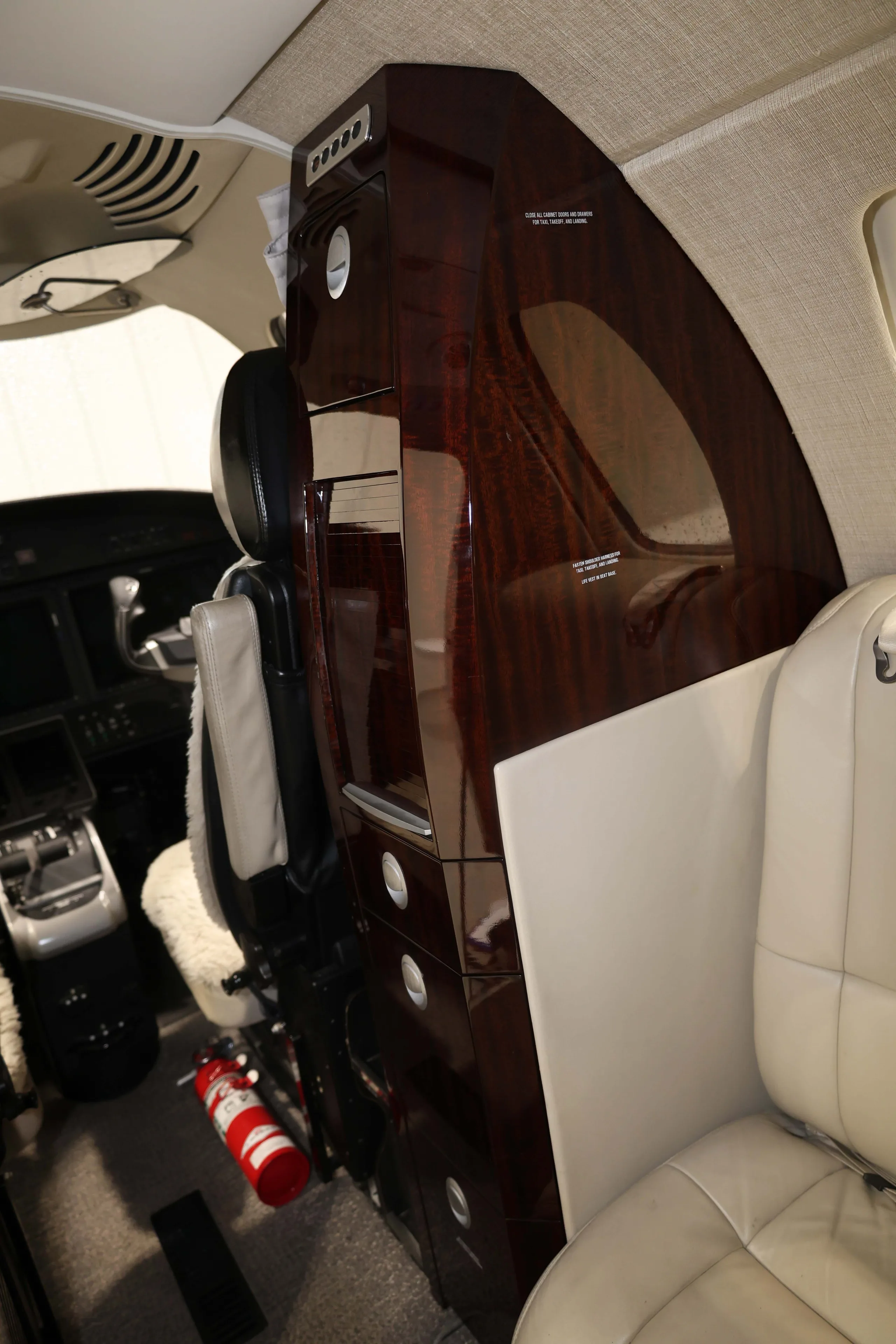 A close-up view of the forward cabin area of a business jet, showing a dark wood cabinet and a fire extinguisher.