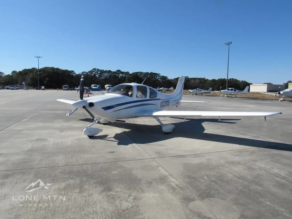 A white Cirrus SR20 aircraft with blue and grey stripes is parked on a concrete ramp, viewed from the front-right.