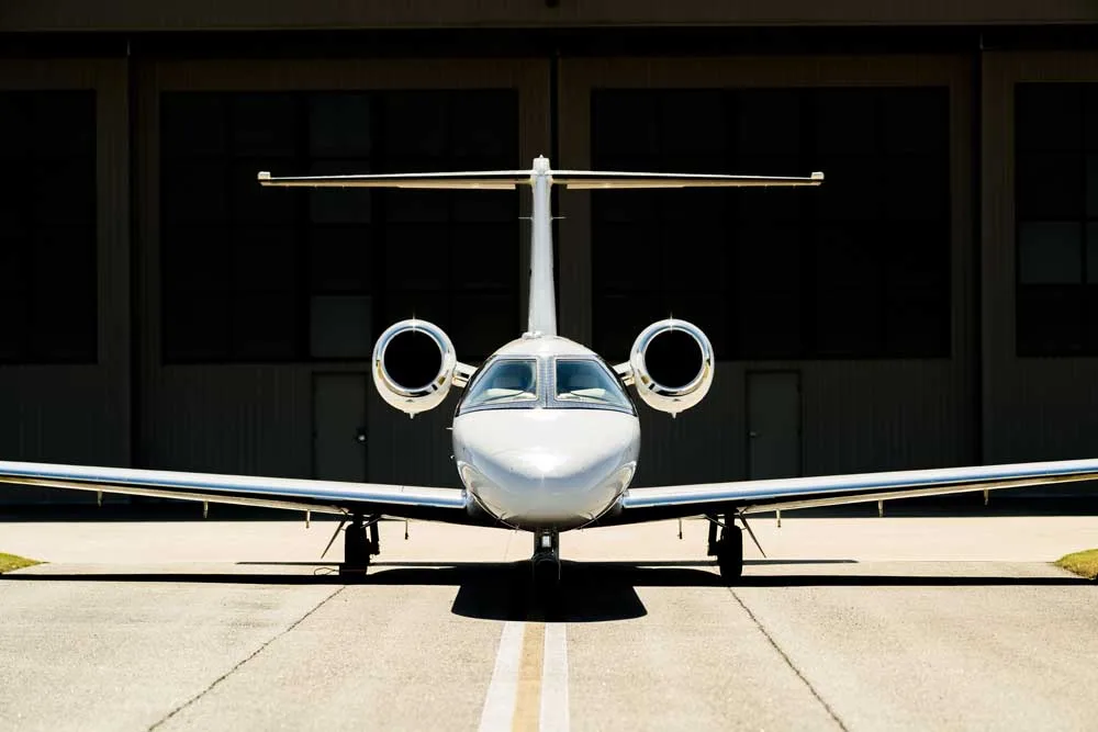 A white business jet is parked head-on on an asphalt tarmac in front of a dark hangar.