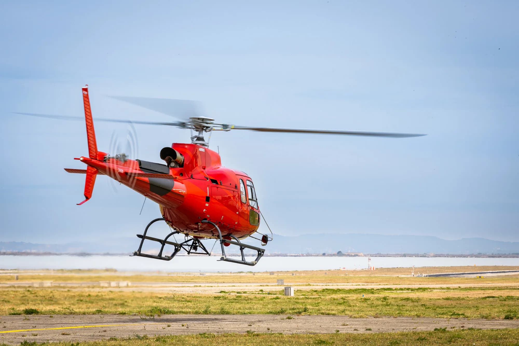 A red Airbus H125 helicopter is captured mid-takeoff from a grassy airfield, with a body of water and distant hills in the background.