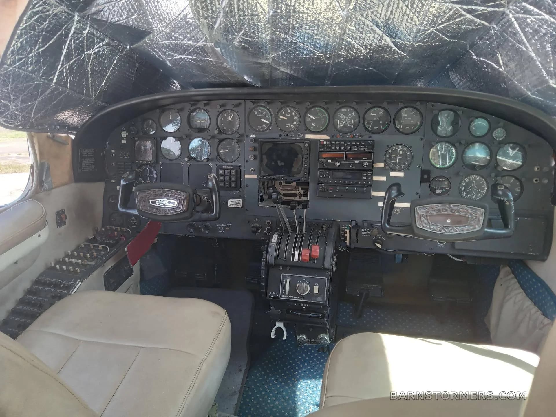 The cockpit of a twin-engine aircraft, featuring a complex instrument panel with analog gauges, two yokes, and a central throttle quadrant, with a sunshade covering the windshield.