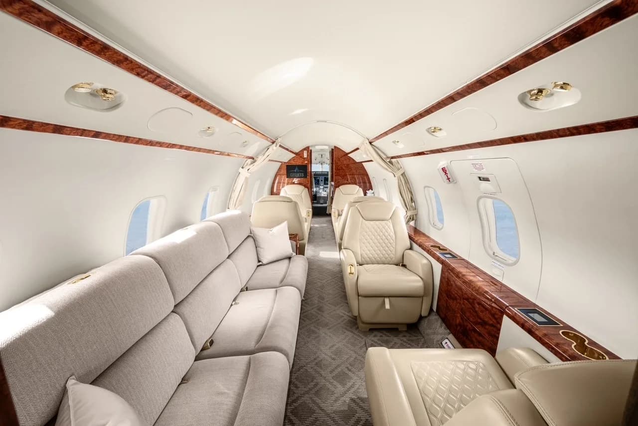 A wide view of a business jet cabin with a grey sofa, cream leather seats, and polished wood paneling.
