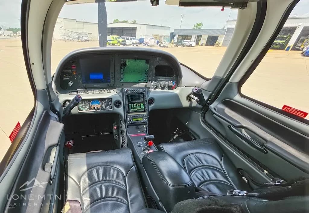 The cockpit of a Cirrus SR22 aircraft, showing the instrument panel and front seats, with an airfield visible through the windshield.