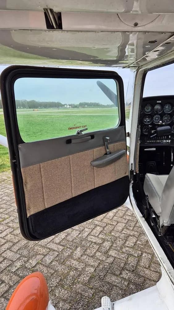 An open aircraft door with a brown fabric panel, showing the cockpit and a grassy airfield outside.
