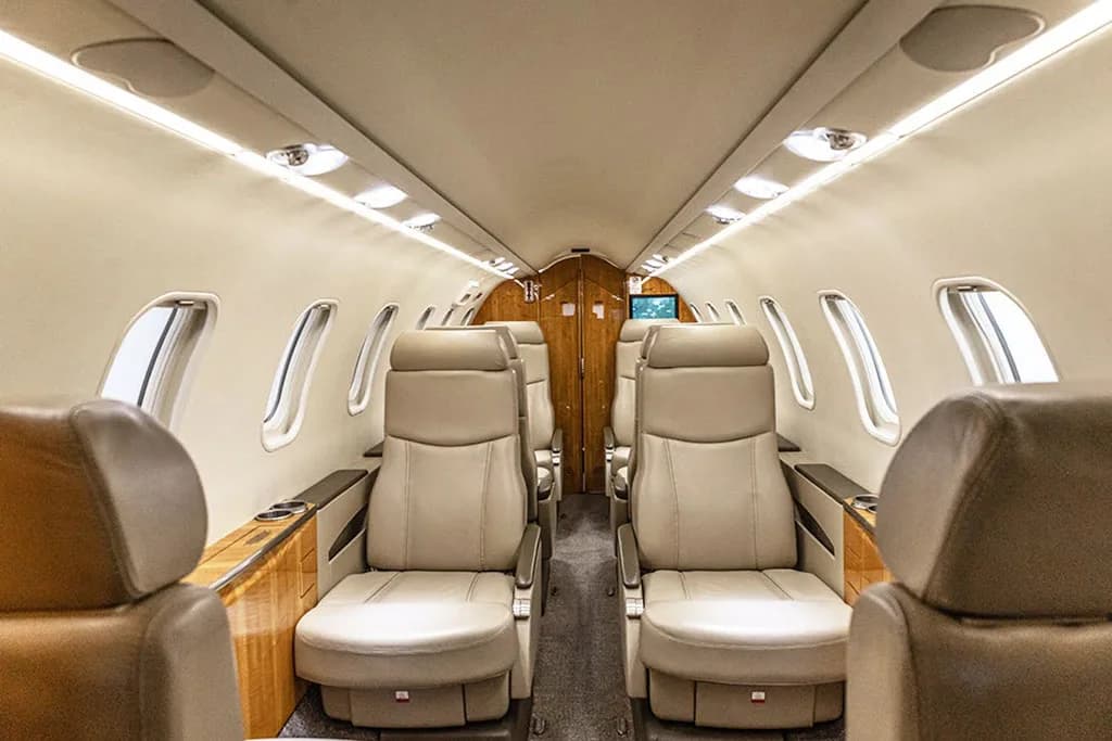 The interior cabin of a private jet with beige leather seats and overhead lighting, looking towards the front.