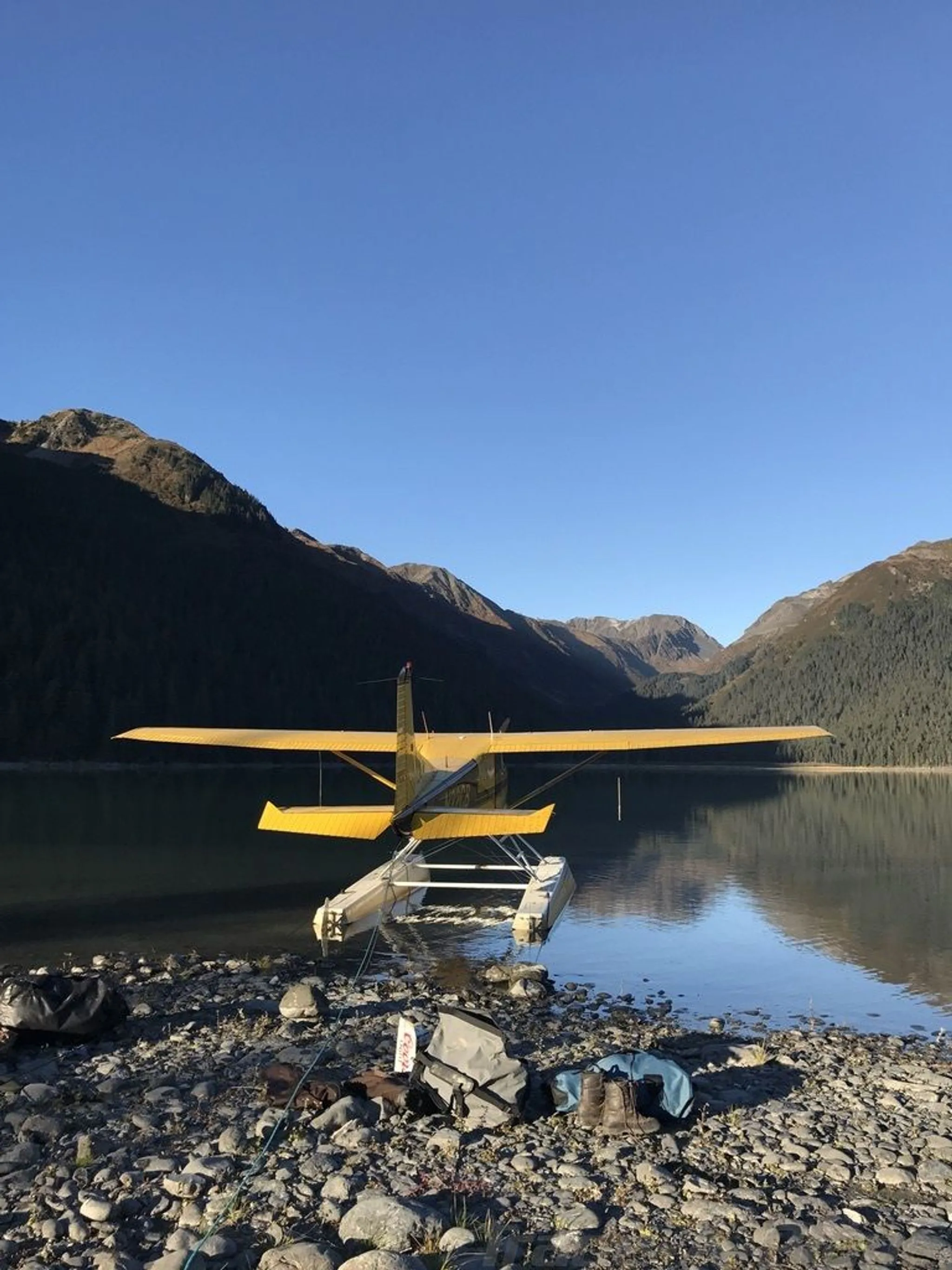 A yellow floatplane is beached on a rocky shore in a mountainous lake setting under a clear blue sky.