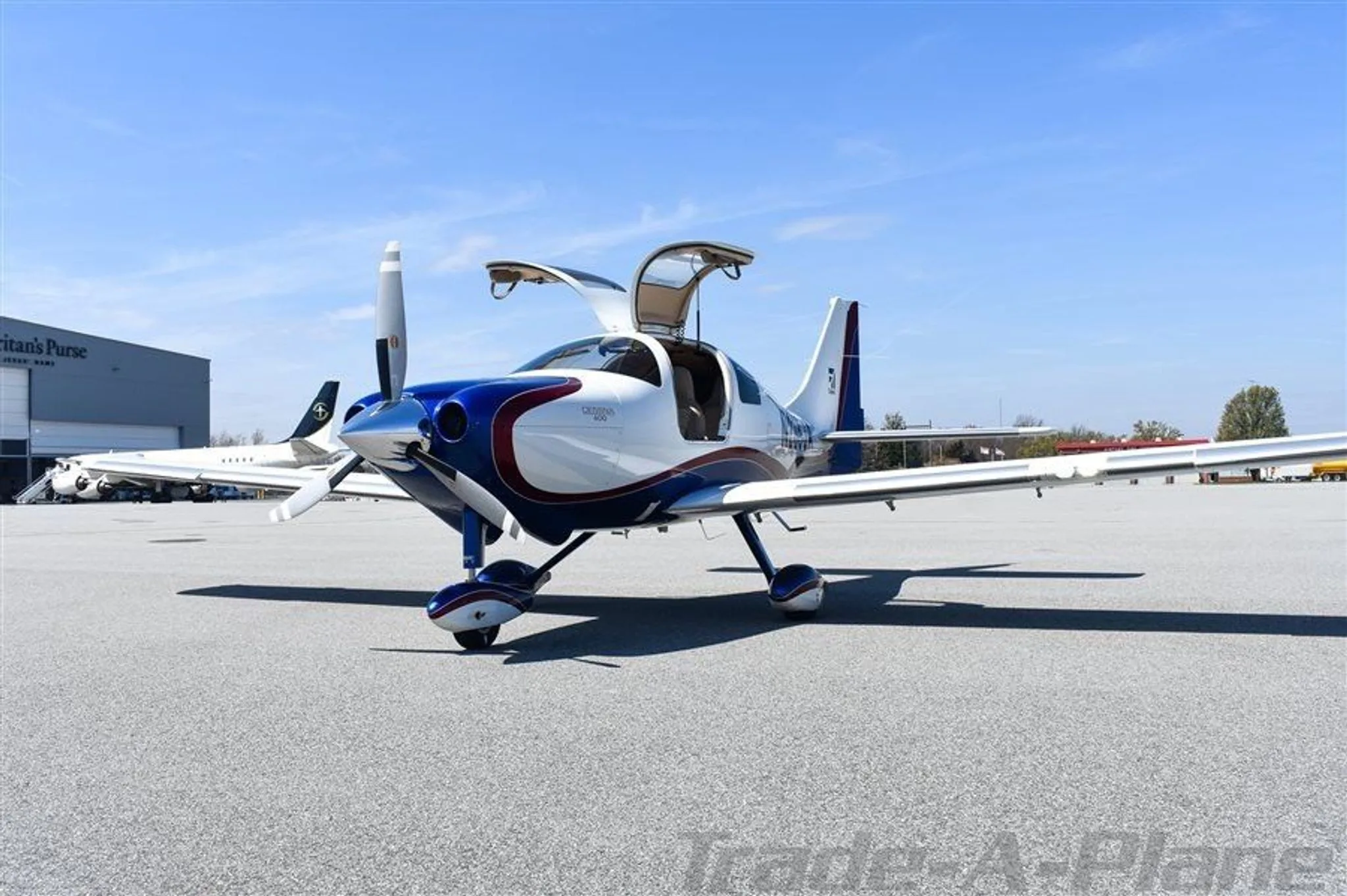 The white, blue, and red Cessna 400 aircraft N235JA is shown with its gull-wing doors open, parked on an airfield.