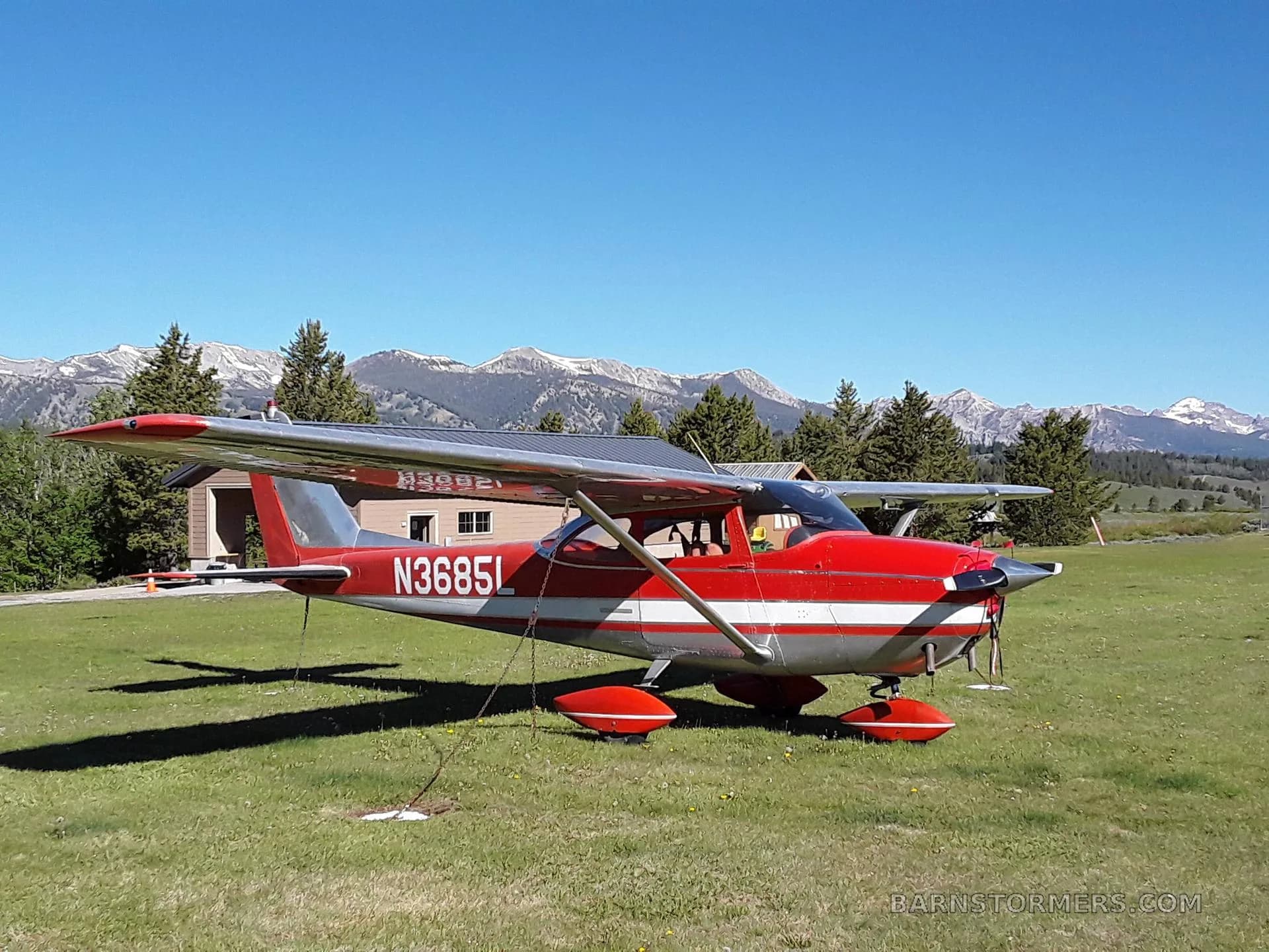 A red and white Cessna 172 (N36851) parked on a grassy field with snow-capped mountains in the background.