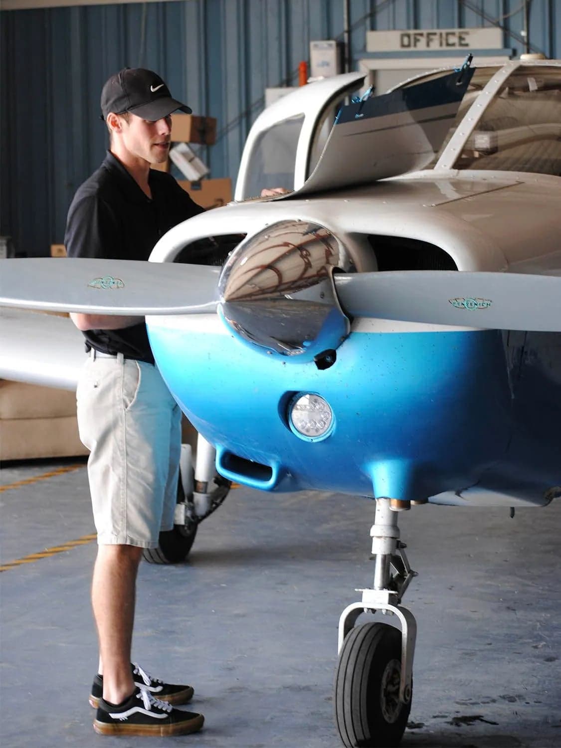 A person in a black cap inspects the engine of a blue and white single-engine aircraft in a hangar.