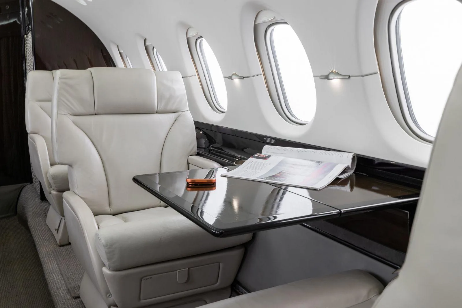 A close-up of a light leather cabin seat with a fold-out table holding a magazine.