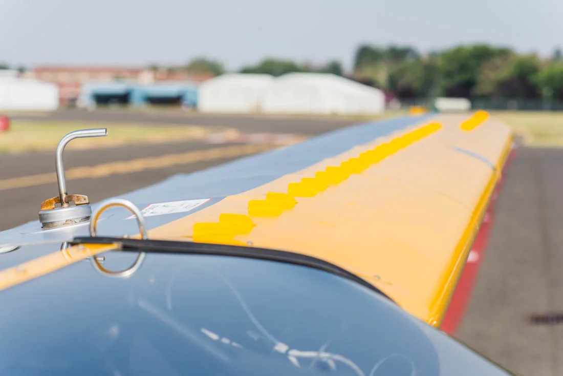A close-up view of the yellow wing of a Carbon Cub aircraft shows the fuel cap and vortex generators.
