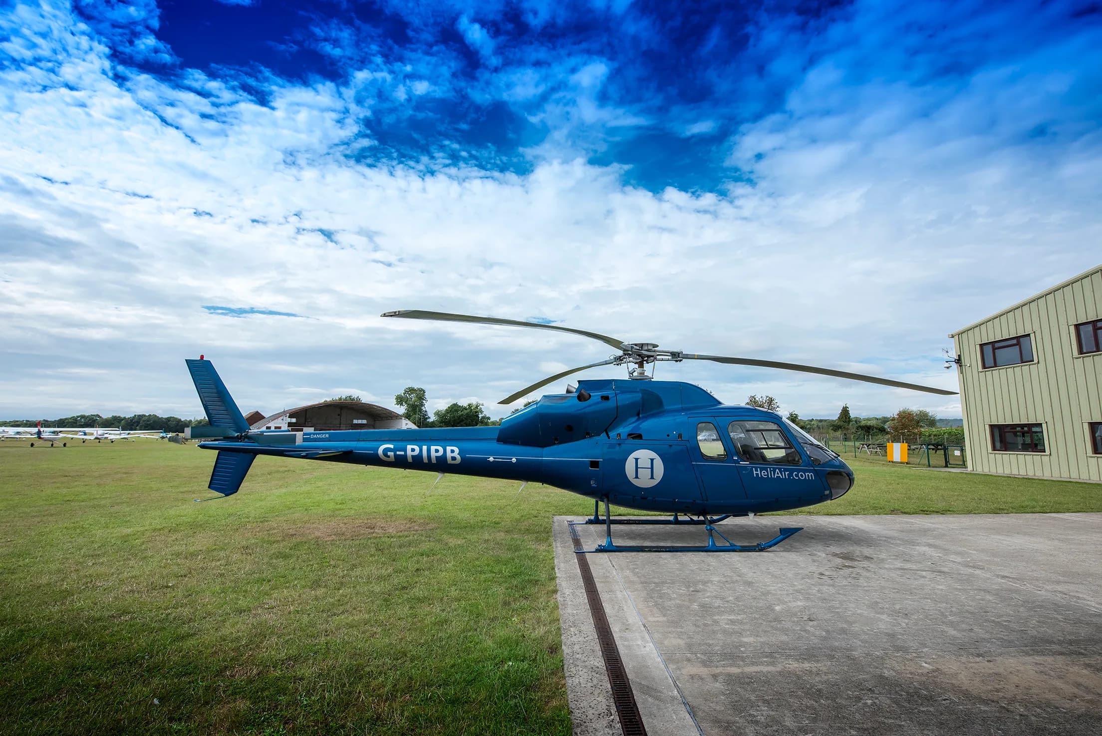 A blue G-PIPB helicopter with 'HeliAir.com' branding is parked next to a building at an airfield under a cloudy sky.