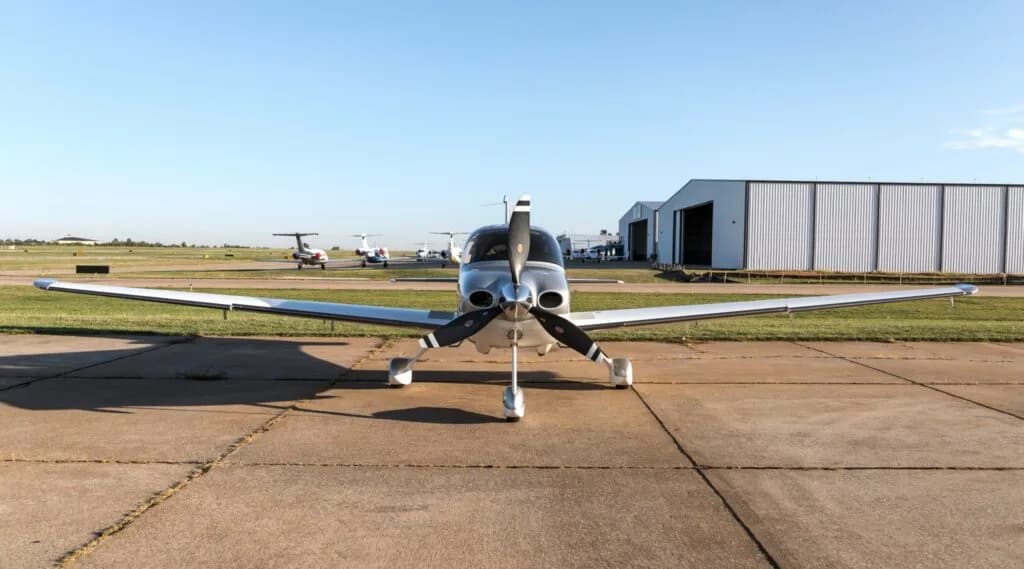 A head-on view of a silver and white Cirrus SR22 G3 Turbo aircraft (N678GW) on an airfield with hangars in the background.