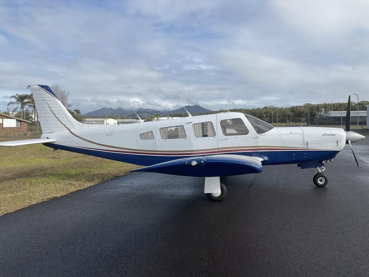A white and blue Piper Saratoga aircraft with brown and gold stripes is parked on a wet tarmac with mountains in the background.