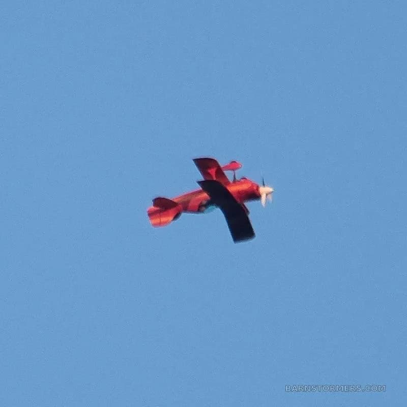 A red biplane with dark wings is seen flying against a clear blue sky.