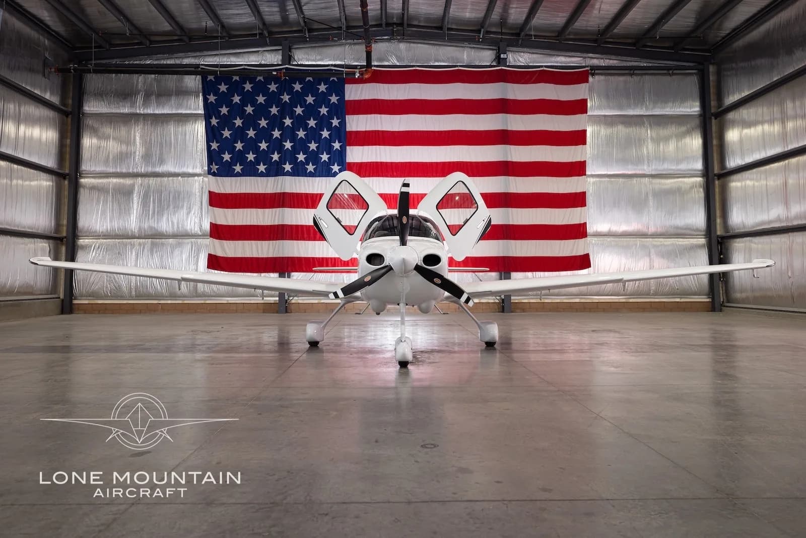 Front view of a white Cirrus SR22 aircraft with registration N327TM, parked in a hangar with an American flag backdrop, with its gull-wing doors open.
