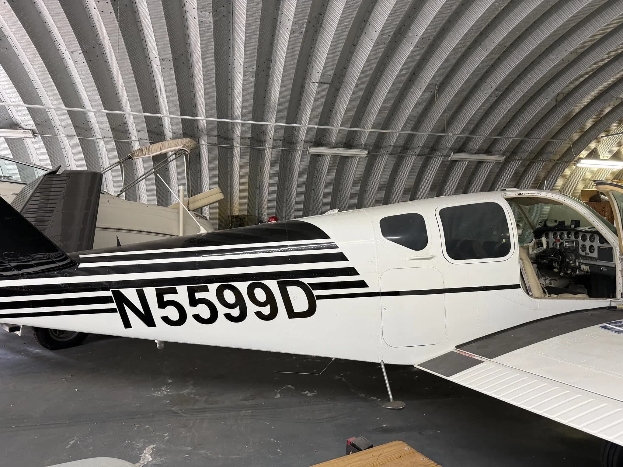 A white single-engine aircraft with black stripes and registration N5599D is parked inside a corrugated metal hangar, with its cockpit door open.