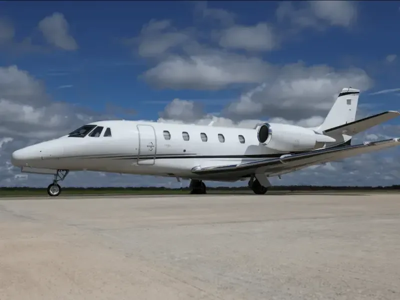A white business jet with black accent stripes parked on a tarmac under a partly cloudy blue sky.
