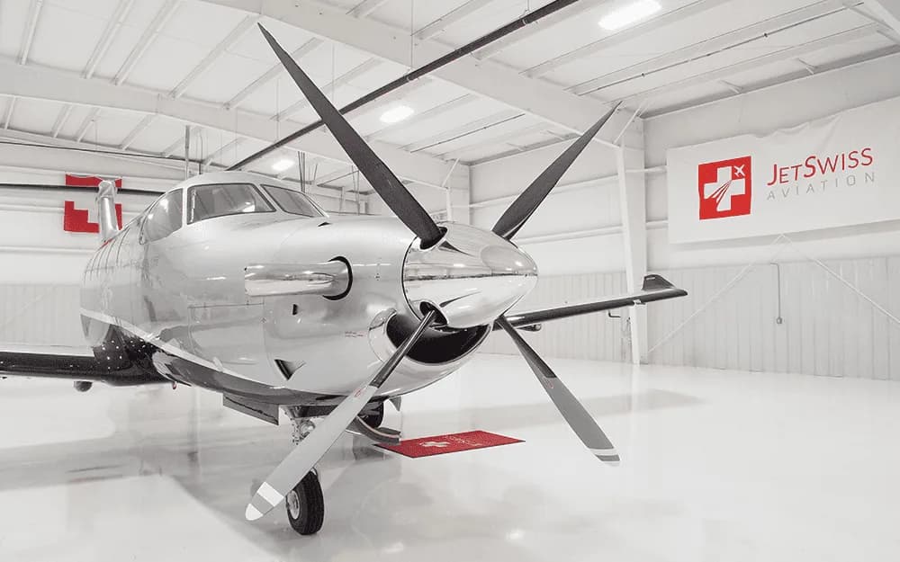 The front view of a silver and black Pilatus PC-12 NG aircraft with its large four-bladed propeller, parked in a hangar.