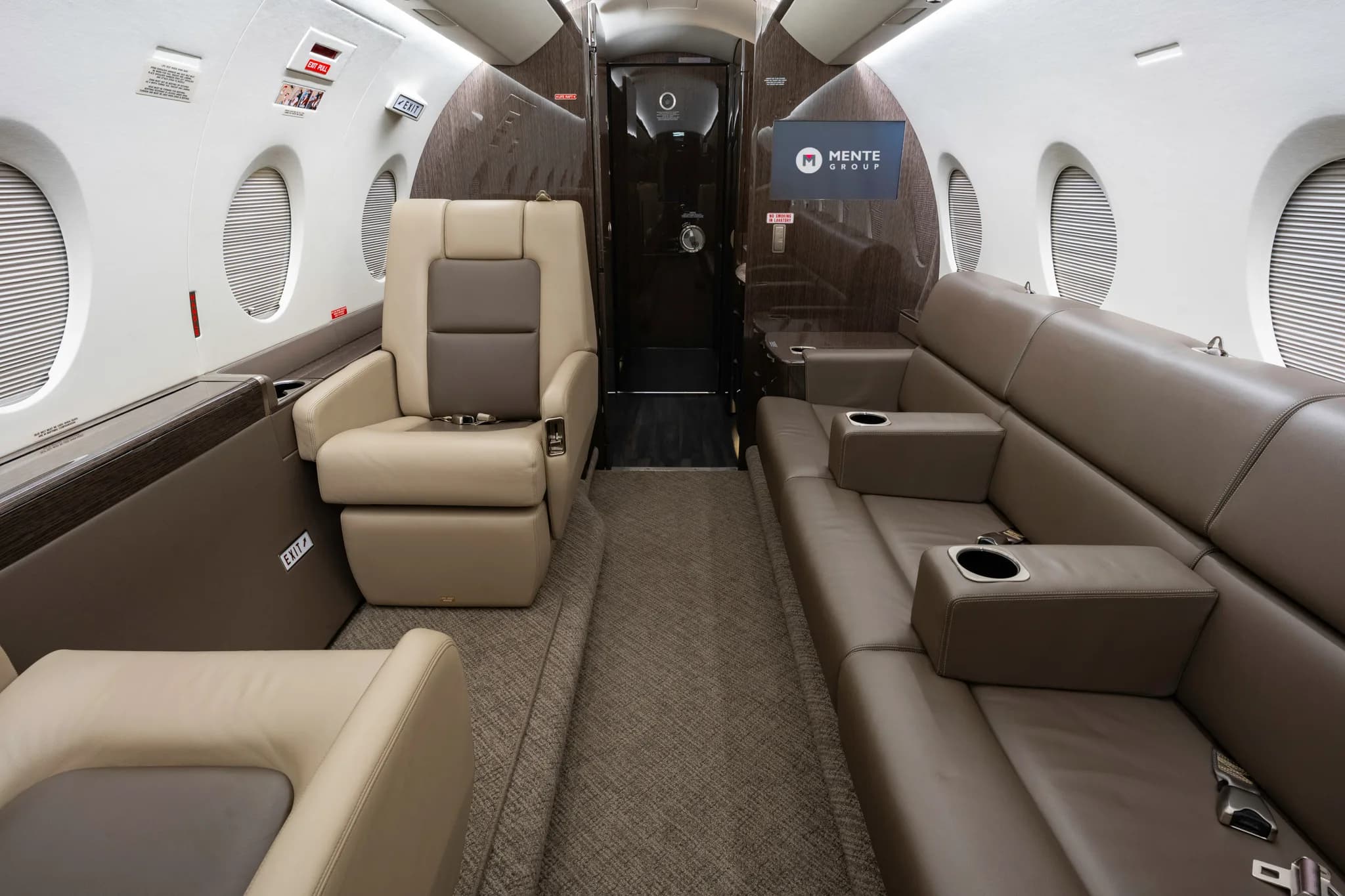The luxurious interior of a private jet cabin, featuring a single beige armchair and a long brown sofa, with a screen displaying a logo at the rear.