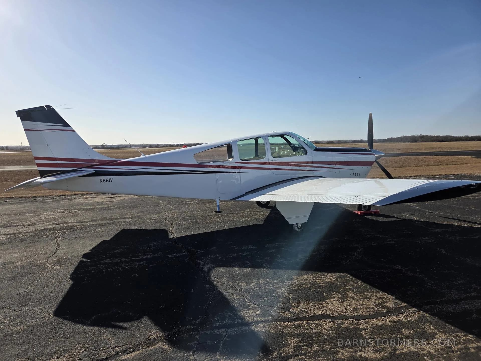 A side view of a white single-engine aircraft with red and black stripes, registration N661V, is parked on an asphalt tarmac under a bright sun.