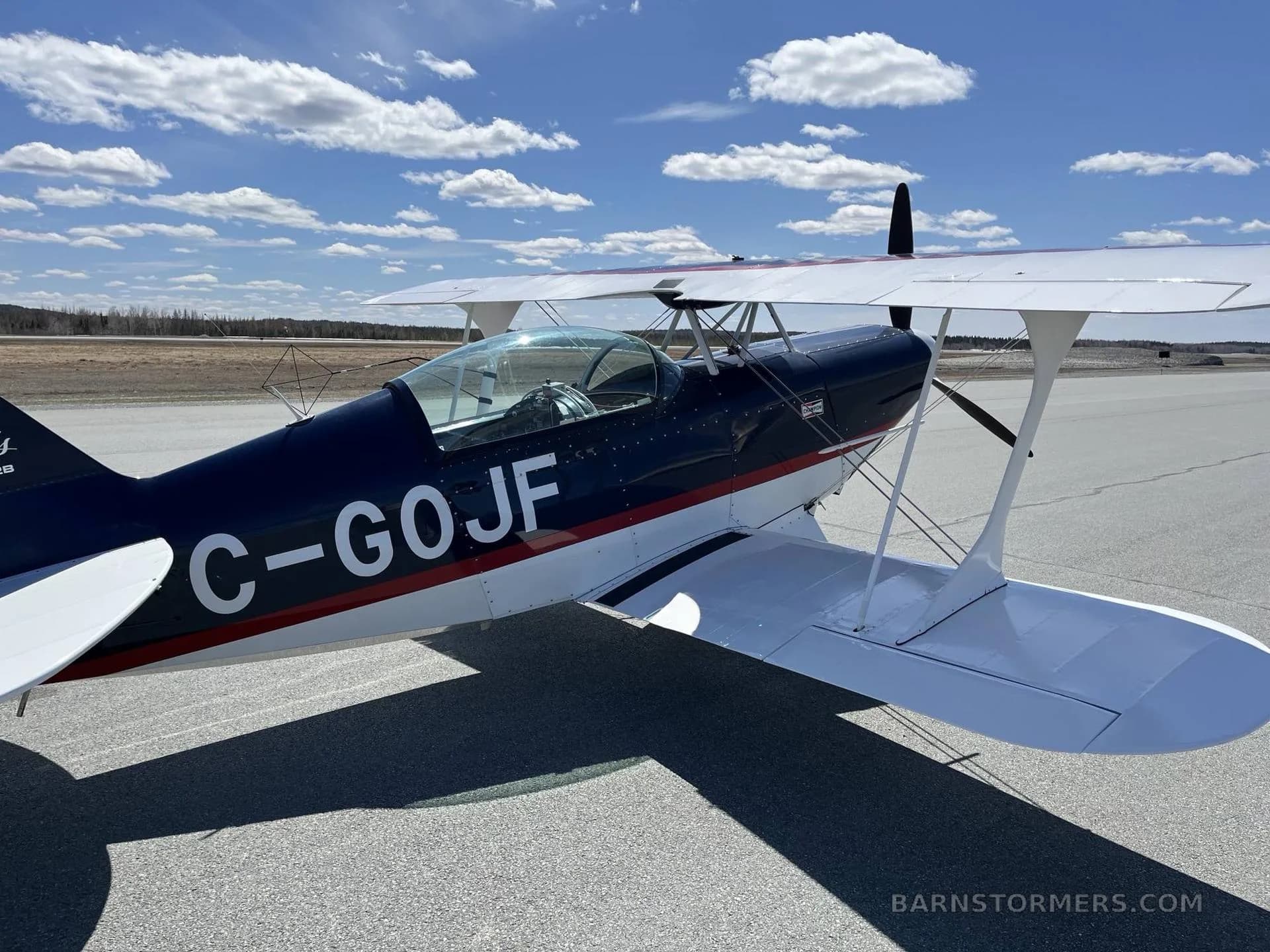 A side view of the dark blue and white biplane C-GOJF parked on an airfield with a blue sky and clouds.