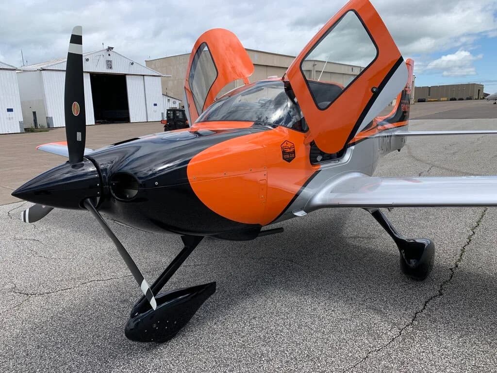 Front view of a Cirrus SR20 aircraft with orange, black, and silver paint scheme, doors open, on an airfield.