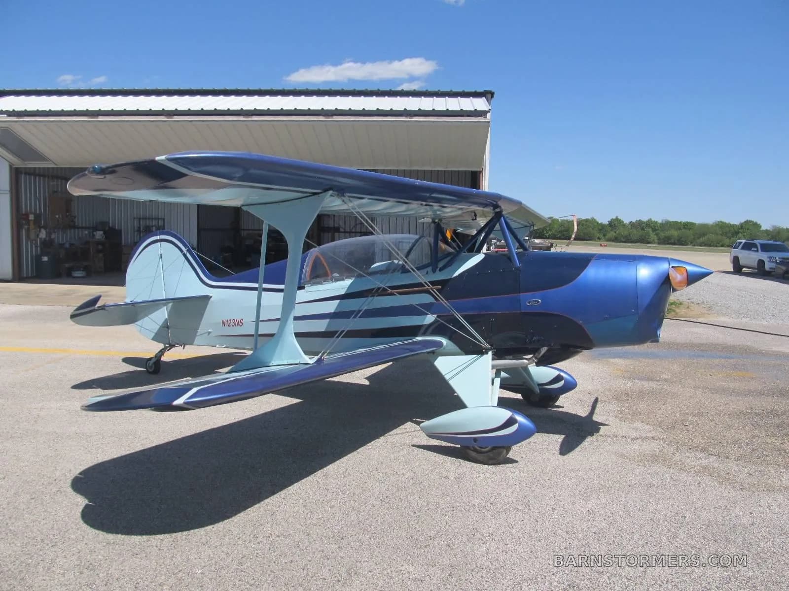 A blue and light blue biplane with an orange propeller spinner is parked on an asphalt apron in front of an open hangar under a blue sky.