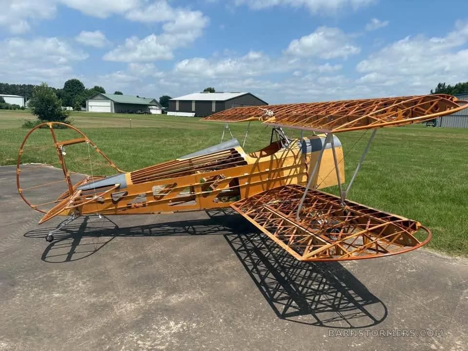 A partially assembled wooden biplane aircraft frame with exposed structure sits on an asphalt pad outdoors.